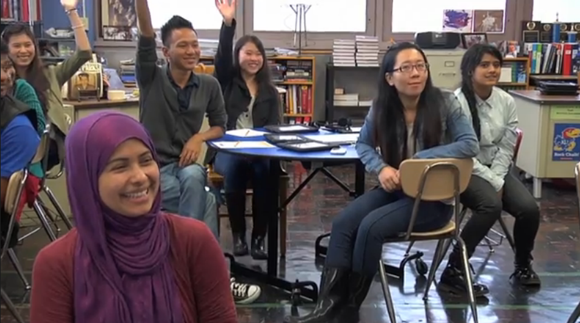 A diverse group of students sits in a classroom facing forward. They are smiling and several have their hands raised.